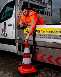 Glimlachende medewerker van Versluys Verkeerstechniek met duim omhoog naast bedrijfsbus en oranje verkeerskegel